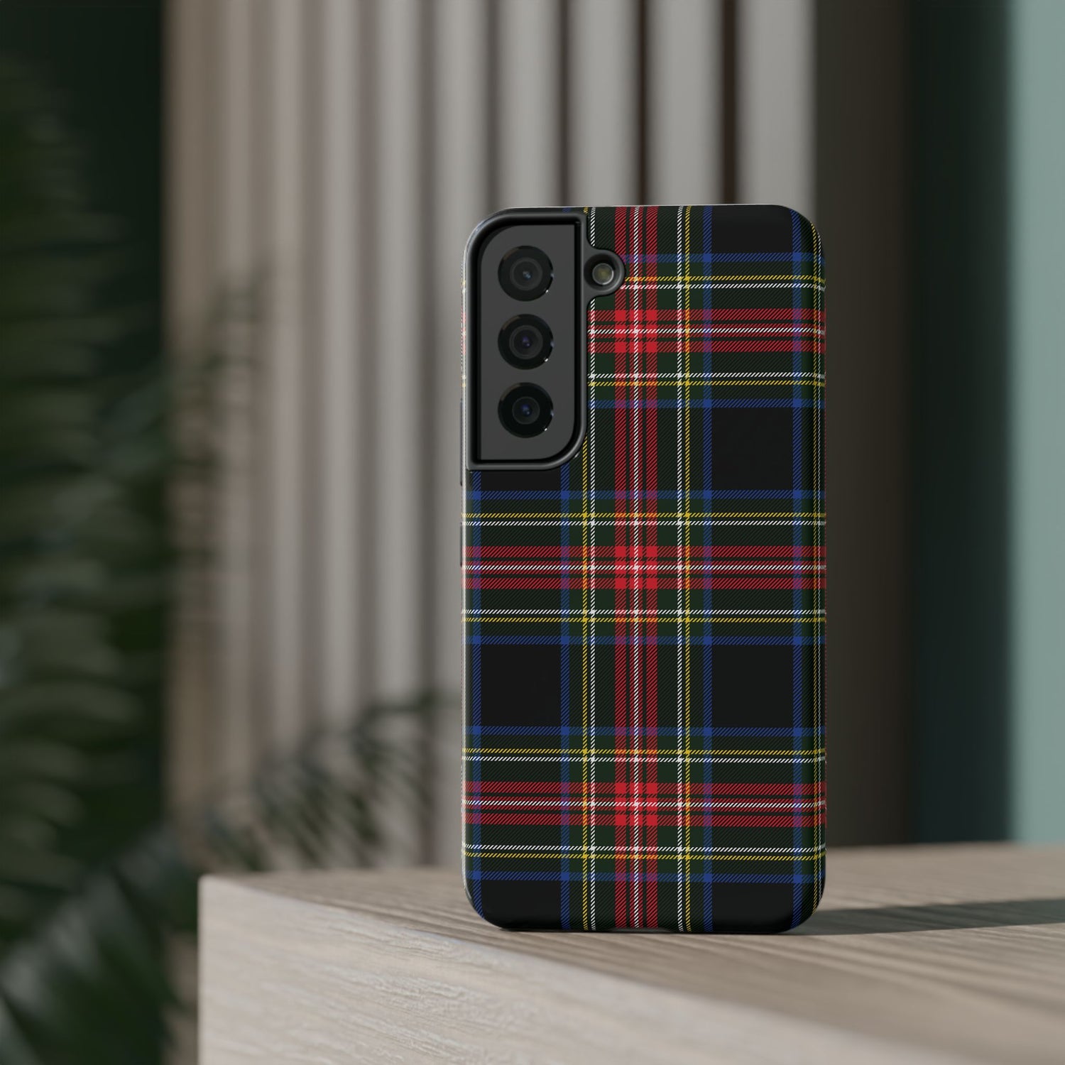 A phone case with a Christmas tartan plaid black pattern, featuring a camera module on the back, placed on a wooden surface with a plant in the background.