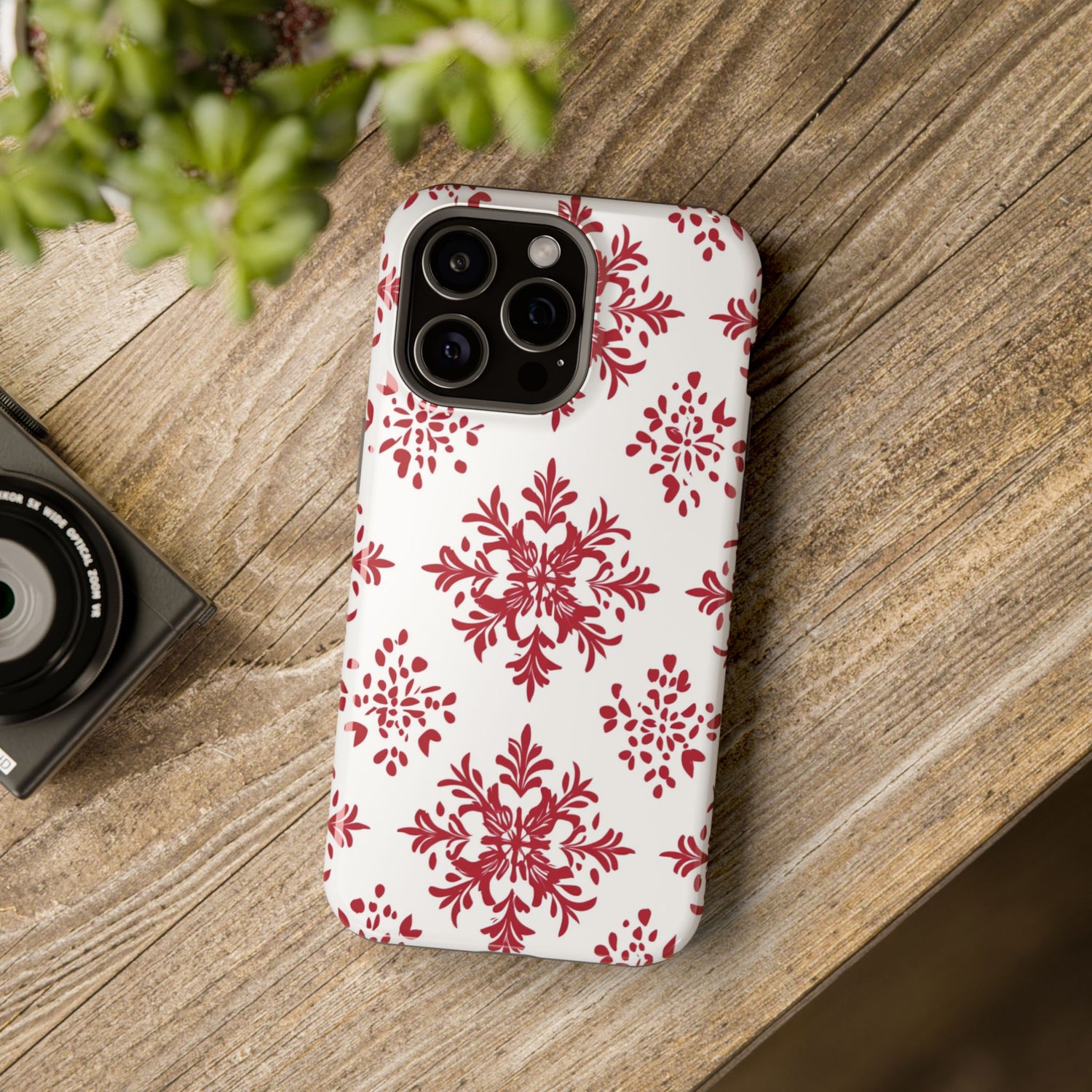 A phone case with a red and white snowflake pattern, featuring a camera cutout on the back, placed on a wooden surface next to a small camera.